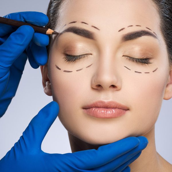 Plastic surgeon drawing dashed lines around closed eyes of girl, above her eyebrow. Hands in blue glove holding pencil and face. Plastic surgery, beauty portrait, closeup
